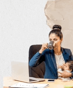 Mujer sentada en una oficina tomando café mientras sostiene a un bebé en brazos, trabajando en una computadora portátil.